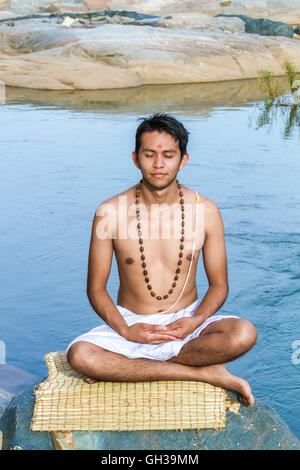 Un giovane sacerdote Indù (bramino), siede in meditazione su una riva di un fiume. Foto Stock