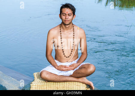 Un giovane sacerdote Indù (bramino), siede in meditazione su una riva di un fiume. Foto Stock