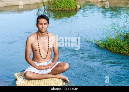 Un giovane sacerdote Indù (bramino), siede in meditazione su una riva di un fiume. Foto Stock