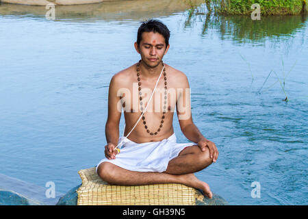 Un giovane sacerdote Indù (bramino) si siede in meditazione su una riva di un fiume. Foto Stock