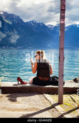 Vista posteriore di una giovane donna seduta sulla riva del lago e applicare il make up, Montreux, Canton Vaud, Svizzera Foto Stock