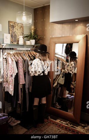 Un 12 anni ragazza indossando un cappello nero in un abbigliamento vintage esplorazione del negozio di vestiti e in piedi accanto a uno specchio Foto Stock