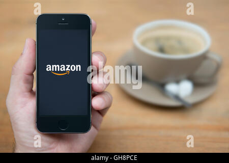 Un uomo guarda al suo iPhone che visualizza il logo Amazon, mentre sat con una tazza di caffè (solo uso editoriale). Foto Stock