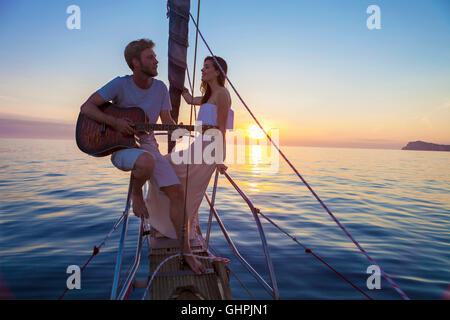 Giovane uomo a suonare la chitarra sulla barca a vela al tramonto Foto Stock