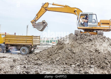 Escavatore giallo è il riempimento di un autocarro con cassone ribaltabile con terreno in corrispondenza del sito di costruzione, il progetto in corso. Foto Stock