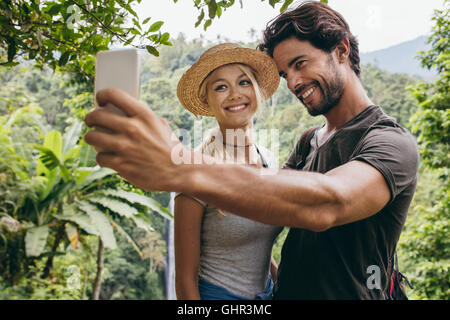 Sorridente coppia giovane tenendo selfie con cascata in background. L uomo e la donna nella foresta tenendo autoritratto con loro cellpho Foto Stock