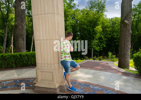 Vista laterale sulla vita contemplativa giovane maschio in piedi vicino al muro di pietra esterno rivolto verso gli alberi nel parco o giardino Foto Stock