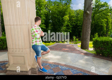 Vista laterale sulla vita contemplativa giovane adulto uomo in piedi vicino al muro di pietra esterno rivolto verso gli alberi nel parco o giardino Foto Stock