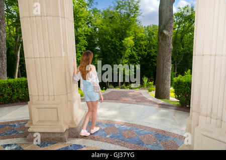 Vista laterale sulla vita contemplativa giovane donna in piedi vicino al muro di pietra esterno rivolto verso gli alberi nel parco o giardino Foto Stock