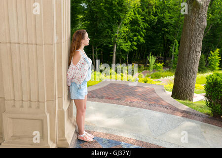 Vista laterale sul contemplativa adulto giovane donna in piedi vicino al muro di pietra esterno rivolto verso gli alberi nel parco o giardino Foto Stock