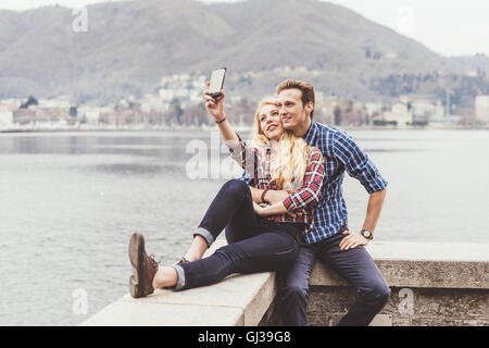 Coppia giovane seduto sulla parete del porto tenendo selfie smartphone, Lago di Como, Italia Foto Stock