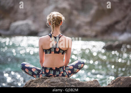 Vista posteriore della giovane donna a praticare yoga lotus posizione sul mare rocce, Maiorca, SPAGNA Foto Stock