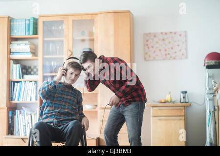 Giovane uomo con sedia a rotelle ascoltando la musica delle cuffie con un amico in cucina Foto Stock