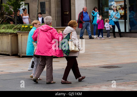 Tre donne anziane indossando variopinti impermeabili felicemente a chiacchierare con loro mentre si cammina insieme nel centro di Dundee, Regno Unito Foto Stock