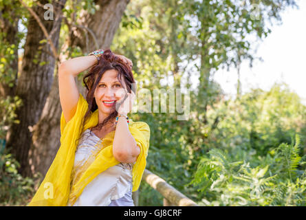 Struttura elegante e affascinante donna matura con scialle giallo e silver top è sorridente in un parco verde durante la riproduzione con i capelli Foto Stock