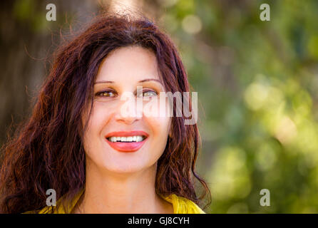 Struttura elegante e affascinante donna matura con abito giallo è sorridente in un parco verde con marrone e rame capelli ondulati Foto Stock