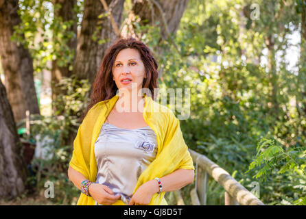 Struttura elegante e affascinante donna matura con mantello giallo e scialle e silver top sta parlando in un parco verde sventolare le mani Foto Stock