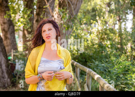 Struttura elegante e affascinante donna matura con mantello giallo e scialle e silver top sta parlando in un parco verde sventolare le mani Foto Stock