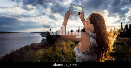 Ragazza giovane con i capelli svolazzanti al vento fotografare digitale compressa bellissimo paesaggio fluviale in serata estiva, donna prendendo photo touch pad fotocamera durante il tempo libero. Vista panoramica. Foto Stock