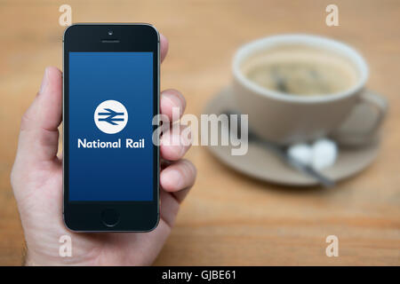 Un uomo guarda al suo iPhone che visualizza il National Rail logo, mentre sat con una tazza di caffè (solo uso editoriale). Foto Stock