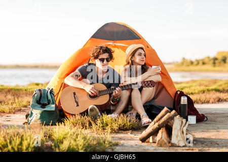 Ritratto di una giovane coppia seduta con chitarra vicino falò in campeggio Foto Stock