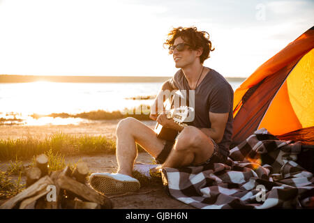 Ricci sorridente giovane uomo seduto vicino alla tenda turistico e a suonare la chitarra Foto Stock