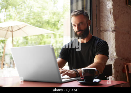 Bello barbuto imprenditore vestita di nero Tshirt Notebook di lavoro tavolo in legno Urban Cafe.giovane manager Notebook di lavoro interni dal design moderno luogo.Coworking del processo di avvio di Business.Filtro di colore. Foto Stock