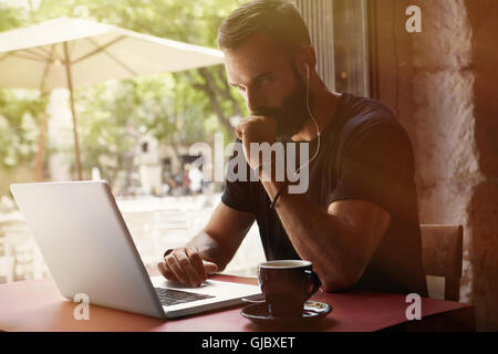 Concentrato giovane imprenditore barbuto vestita di nero Tshirt Notebook di lavoro Urban Cafe.uomo seduto tabella tazza di caffè ascoltando musica.Coworking del processo di avvio di Business.sfondo sfocato.Filtro di colore. Foto Stock