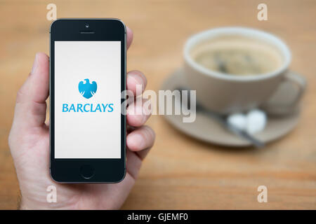 Un uomo guarda al suo iPhone che visualizza la Barclays Bank logo, mentre sat con una tazza di caffè (solo uso editoriale). Foto Stock