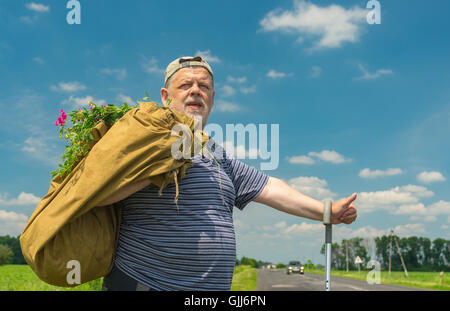 Senior uomo con sacco e mazzo di fiori selvatici in piedi su una strada e che mostra segni di Hitchhiker Foto Stock