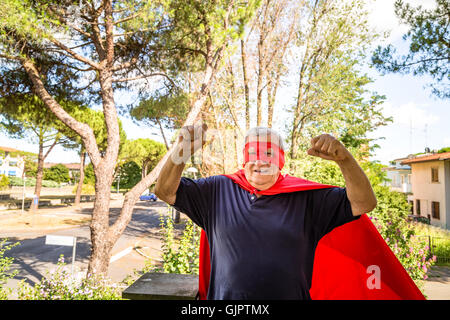 Divertente e sorridente uomo senior in posa come supereroe con red cape e maschera è che mostra i muscoli dei bracci di sollevamento in un tranquillo quartiere residenziale Foto Stock