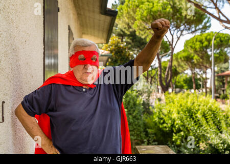 Divertente e felice uomo senior in posa come supereroe con red cape e maschera è salita del braccio si prepara a volare in un tranquillo quartiere residenziale Foto Stock