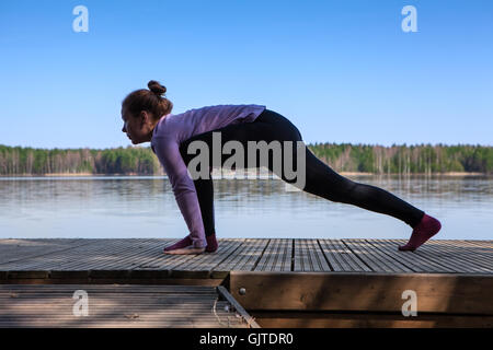 La donna caucasica rendendo semplice lo Yoga asana sul molo in legno sulla riva del lago Foto Stock