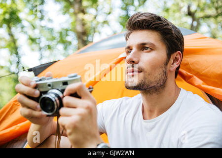 Concentrato giovane con foto vecchia telecamera seduta vicino tenda turistico nella foresta Foto Stock