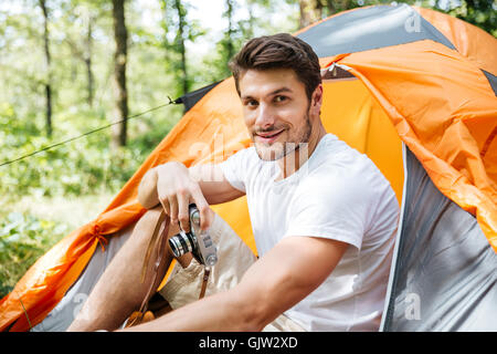 Sorridente giovane uomo seduto in tenda turistico e usando la vecchia foto fotocamera nella foresta Foto Stock