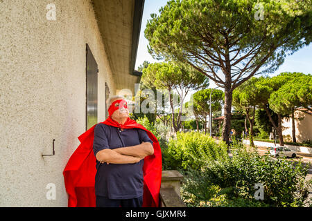 Divertente e sorridente senior uomo vestito da supereroe con mantello rosso e la maschera è in piedi con le braccia incrociate sul balcone della sua casa in un tranquillo quartiere residenziale Foto Stock