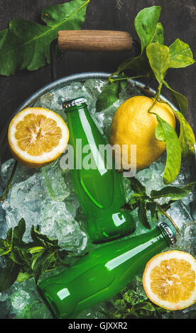 Bottiglie di limonata verde su scaglie di ghiaccio nel vassoio di metallo con limoni freschi e menta su legno scuro sfondo, vista dall'alto, vertica Foto Stock