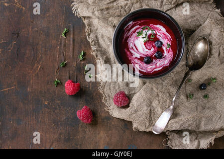 Nero vaso di ceramica di lampone dolce zuppa con crema, servito con il cucchiaio, frutti di bosco freschi e timo su un letto di sacco su legno vecchio t Foto Stock