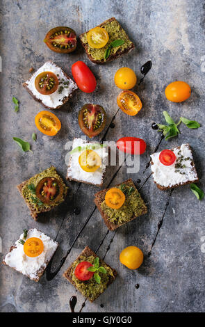 Varietà di piccoli panini di segale con colorati di pomodori ciliegini, ricotta e pesto, servita con erbe fresche e balsa Foto Stock