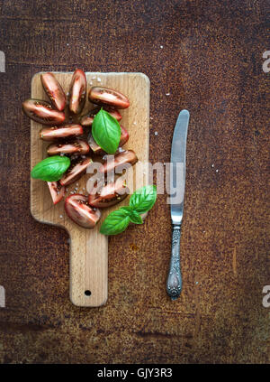 Freschi fette di pomodoro con il sale e le foglie di basilico sul tagliere di legno su metallo grunge sfondo arrugginito, vista dall'alto Foto Stock