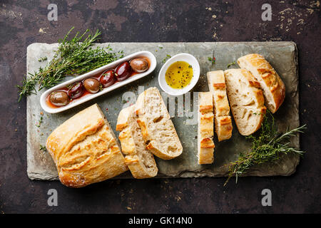 La ciabatta fresca pane tagliato a fette su pietra ardesia bordo con olive, olio e erbe su sfondo scuro Foto Stock