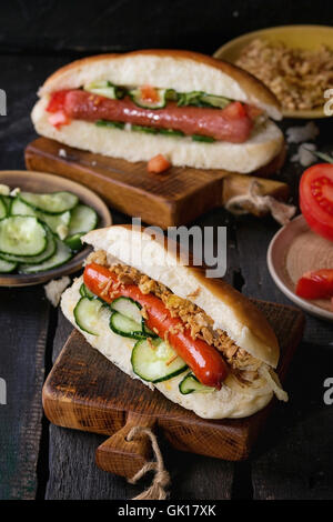 Due fatti in casa di hot dog con salsiccia, cipolla fritti, pomodori e cetrioli, servita con ingredienti in diverse piastre su legno c Foto Stock