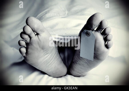 Primo piano dei piedi di un corpo morto coperta con un foglio con un tag vuoto legato sul dito grosso del suo piede sinistro, in monocromia, w Foto Stock