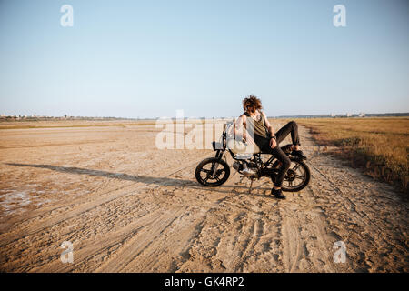 Giovane uomo brutale recante sulla sua moto nel deserto e posa Foto Stock