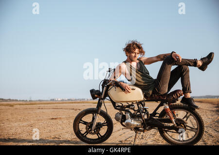 Giovane uomo brutale recante sulla sua moto nel deserto e posa Foto Stock