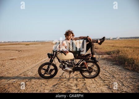 Giovane uomo brutale recante sulla sua moto nel deserto e posa Foto Stock