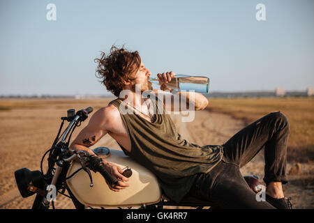 Giovane uomo brutale recante sulla sua moto nel deserto e acqua potabile Foto Stock