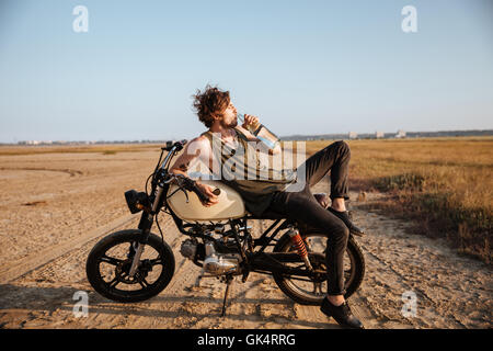 Giovane uomo brutale recante sulla sua moto nel deserto e acqua potabile Foto Stock