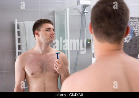 Scontento giovane con tagli sul suo volto dopo la cattiva tenuta di rasatura del rasoio mentre in piedi di fronte allo specchio nel moderno bagno rivestito di piastrelle a casa. Foto Stock