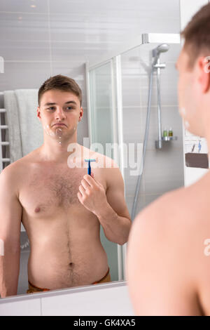 Bel giovane maschio con tagli sul suo volto dopo la cattiva tenuta di rasatura del rasoio mentre in piedi di fronte allo specchio nel moderno bagno rivestito di piastrelle a casa. Foto Stock